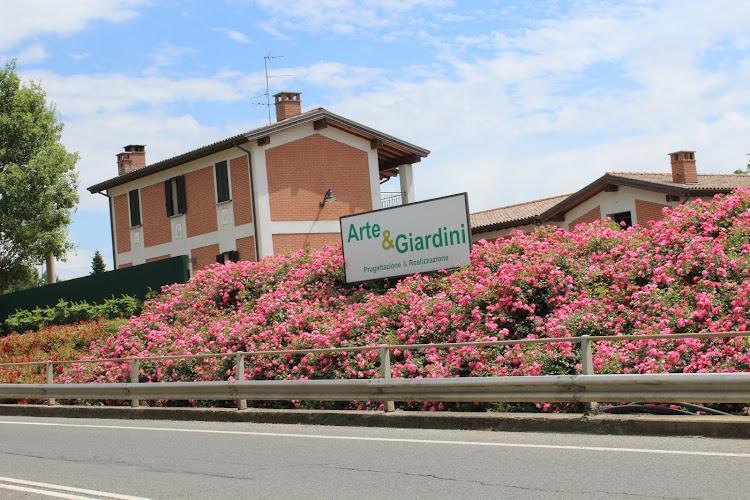 Dietro un rigoglioso giardino pieno di fiori rosa si trova un edificio in mattoni a due piani, con un cartello che recita "Arte & Giardini Progettazione | Realizzazione" lungo la ringhiera della strada.