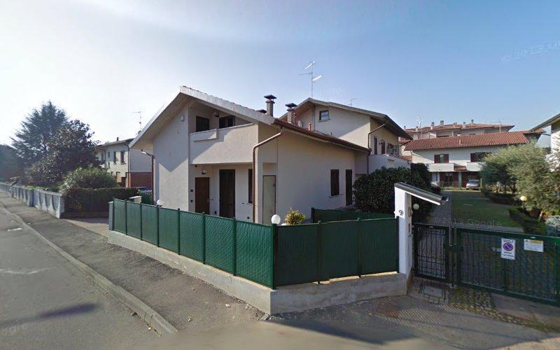 Una casa bianca a due piani con un cortile recintato verde si trova all'angolo di una strada residenziale. Altre case sono visibili sullo sfondo. Il cielo è limpido e la scena è illuminata dal sole.