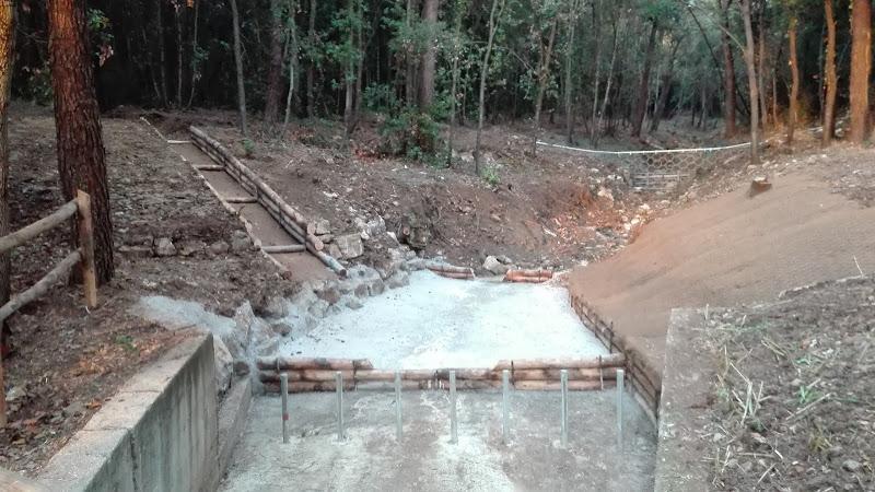 Una struttura di drenaggio o un ponte in cemento in costruzione in una zona boscosa, con ringhiere in legno e un sentiero di ghiaia che vi conduce.