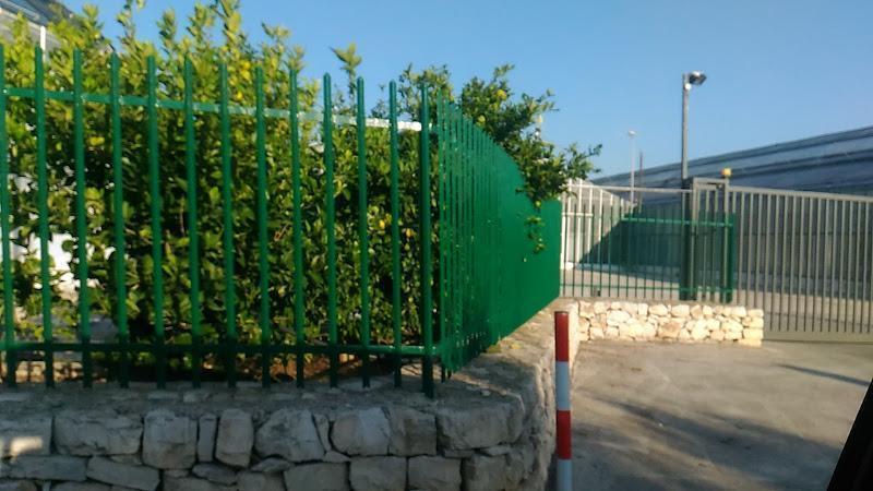 Una recinzione metallica verde racchiude una piccola area di giardino con cespugli, costruita su un muro di pietra. Nelle vicinanze, si vede un palo a strisce accanto all'ingresso di un'area industriale con grandi strutture metalliche.