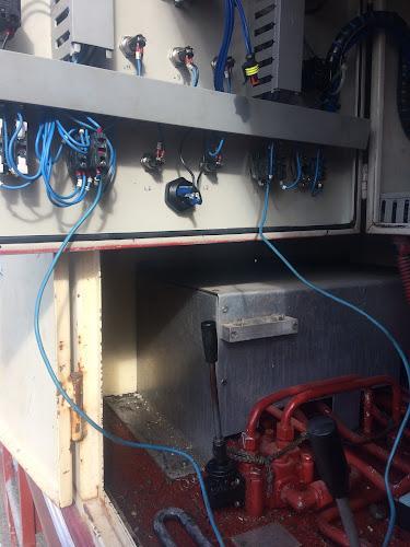 Un pannello di controllo industriale parzialmente aperto con fili blu, pulsanti e leve collegati a vari componenti, che mostra parti meccaniche e tubi all'interno di un involucro di macchinari.