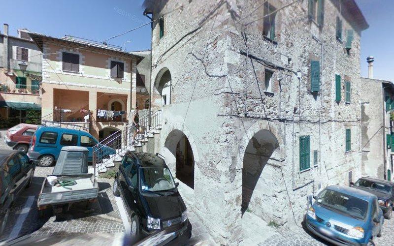Un vecchio edificio in pietra con le persiane verdi si trova all'incrocio di una strada, circondato da auto parcheggiate in un villaggio italiano.