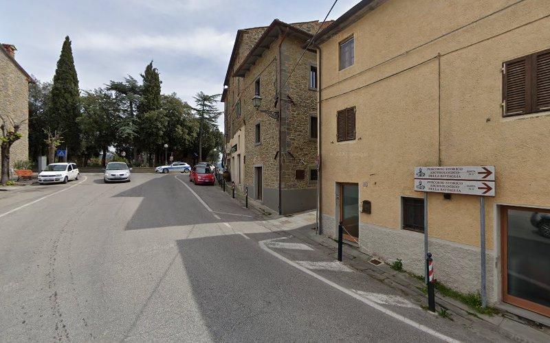 Una strada stretta in una cittadina con edifici beige e in pietra. Ci sono alcune auto parcheggiate e un cartello stradale che indica varie località. Alberi e vegetazione sono visibili sullo sfondo.