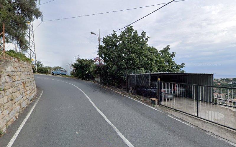 Una strada tortuosa sale in salita accanto a un muro di pietra e una recinzione metallica che racchiude una piccola area di parcheggio con auto. Un veicolo blu è parcheggiato vicino al bordo della strada. Sono visibili alberi e linee elettriche.