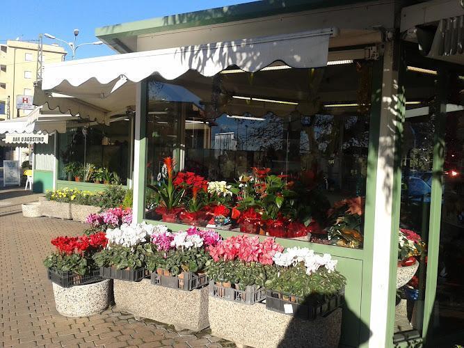 Un negozio di fiori con una tenda verde espone vari fiori e piante colorati in vasi all'esterno. Il negozio si trova su un'area pavimentata con edifici residenziali sullo sfondo.