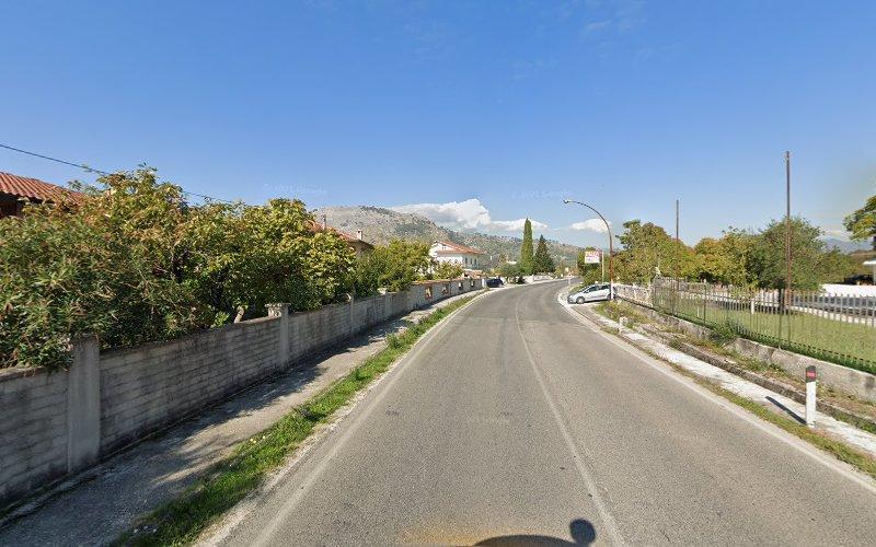 Una strada curva fiancheggiata da muri in pietra e case sulla sinistra, e un'area erbosa con una recinzione sulla destra. Le montagne sono visibili in lontananza sotto un cielo azzurro e terso.