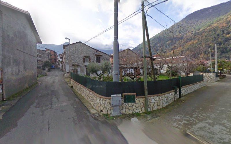 Una strada stretta in un villaggio con case in pietra, linee elettriche e montagne sullo sfondo in una giornata nuvolosa.