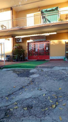 Una vetrina con un ingresso rosso, una tenda da sole e piante vicino alla porta. Un tappeto verde si estende dalla porta fino al marciapiede, che è screpolato e cosparso di foglie.