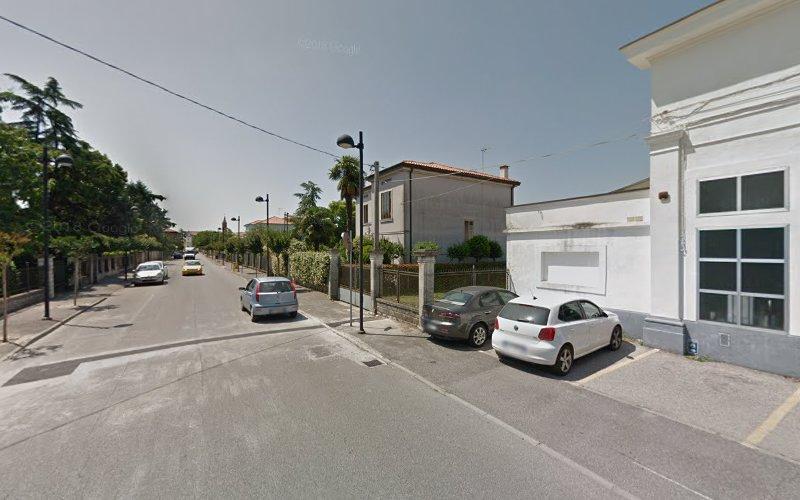 Una strada residenziale con auto parcheggiate lungo i lati, fiancheggiata da case, alberi e lampioni sotto un cielo limpido. Un edificio bianco è sulla destra con proprietà recintate sulla sinistra.
