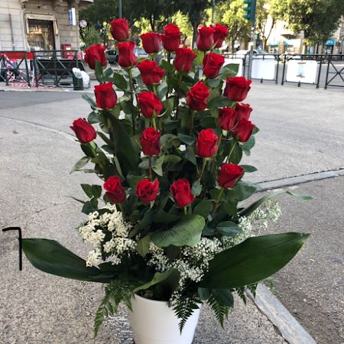 Un vaso bianco pieno di rose rosse e foglie verdi è posto su un marciapiede con una strada e degli edifici sullo sfondo.