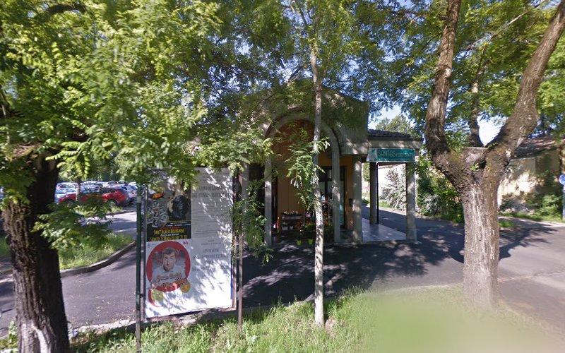 Un edificio con un ingresso ad arco è parzialmente nascosto dagli alberi. Un cartello con testo e immagini è visibile di fronte all'edificio. Un parcheggio con auto è visibile sulla sinistra.