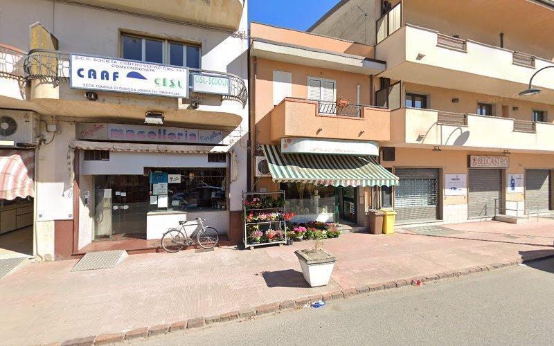 Vista stradale di una fila di piccole attività commerciali, tra cui un bar, una macelleria e un negozio di fiori con una tenda a strisce verdi e bianche e un'esposizione di fiori all'esterno. Una bicicletta è parcheggiata di fronte.