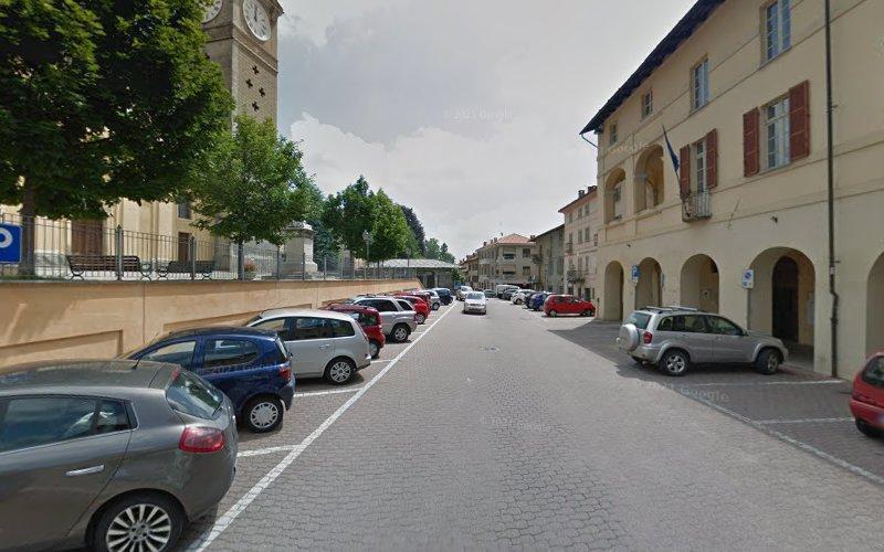 Una strada fiancheggiata da auto parcheggiate su entrambi i lati, accanto a edifici beige con balconi e una torre dell'orologio sullo sfondo in una giornata nuvolosa.