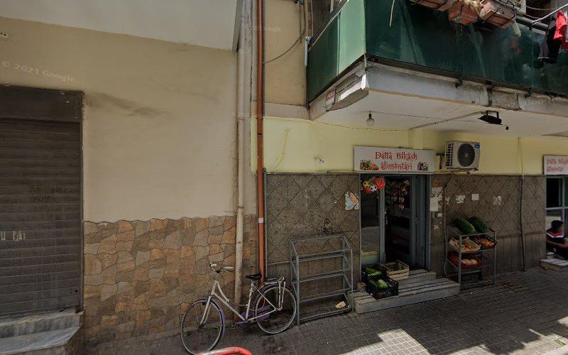 Un piccolo negozio di alimentari all'angolo di una strada con una tenda da sole, una bicicletta parcheggiata fuori e prodotti in casse visibili all'ingresso. Accanto c'è una vetrina chiusa con una saracinesca.