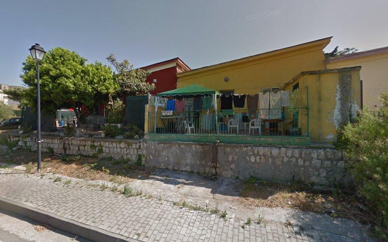Una casa gialla con i panni stesi ad asciugare su una recinzione metallica verde, circondata dal verde e da un muro di pietra, accanto a una strada in una giornata di sole.