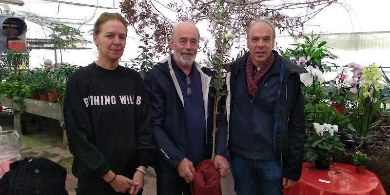 Tre persone sono in piedi insieme in un vivaio, circondate da varie piante in vaso e fiori. La persona al centro tiene in mano un piccolo albero.