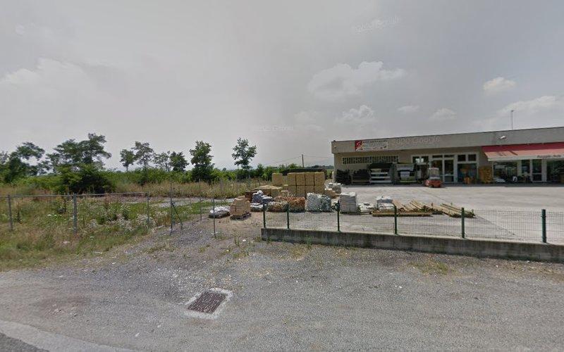 Scena esterna di un deposito di rifornimenti con vari materiali edili e pallet di fronte a un magazzino. L'area è recintata, con alberi e vegetazione sullo sfondo sotto un cielo parzialmente nuvoloso.