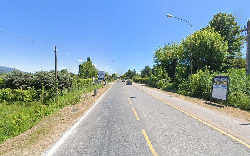 Una strada dritta a due corsie fiancheggiata dal verde sotto un cielo azzurro e terso, con un'auto che guida in lontananza e lampioni che costeggiano i lati. Ci sono cartelli informativi e cartelloni pubblicitari lungo la strada.