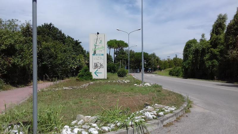 Vista stradale di una strada con una rotonda caratterizzata da un cartello alto con graffiti, paesaggio con rocce e arbusti, lampioni e una pista ciclabile sulla sinistra. Alberi e arbusti costeggiano la strada.