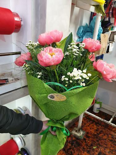 Una persona tiene in mano un bouquet avvolto in carta verde, contenente peonie rosa e fiori bianchi di gypsophila. Lo sfondo include alcuni oggetti rossi e bianchi e oggetti appesi.