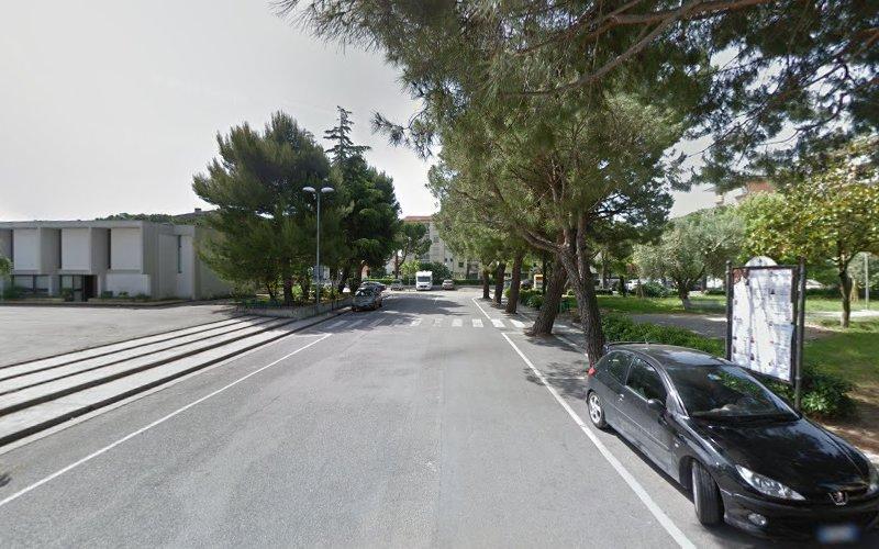 Una strada fiancheggiata da alberi, con un attraversamento pedonale, un'auto nera parcheggiata e un edificio sulla sinistra. C'è un grande cartello sulla destra. Il cielo è limpido.