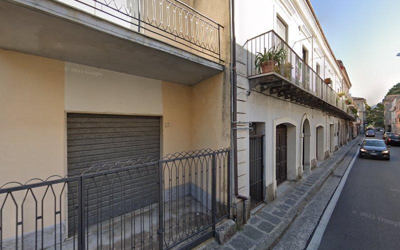 Una strada stretta fiancheggiata da edifici residenziali, con balconi con piante. Sulla strada sono visibili diverse auto parcheggiate e un'auto in movimento.