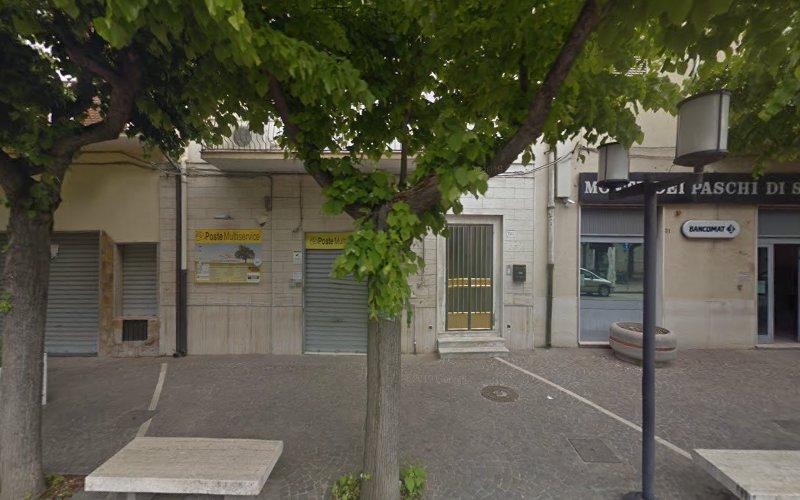 Vista stradale di un edificio con vetrine che mostrano un cartello giallo con la scritta "Poste Italiane", circondato da alberi e persiane chiuse. Un altro cartello recita "Monte dei Paschi di S." Sono visibili delle panchine.
