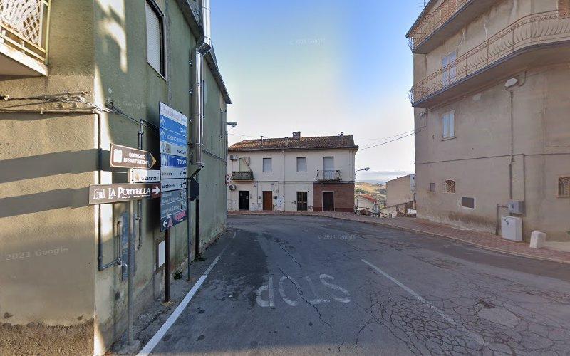 Una strada stretta, leggermente in pendenza, fiancheggiata da edifici con cartelli che indicano le direzioni. Un segnale "STOP" è visibile sul marciapiede screpolato e il cielo è limpido.