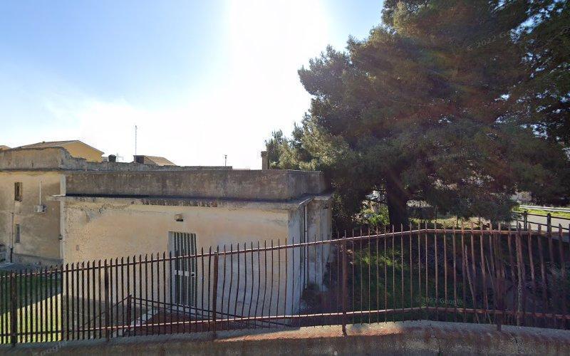 Un piccolo edificio a un piano con tetto piano è circondato da una recinzione metallica. Ci sono grandi alberi e cieli limpidi sullo sfondo.
