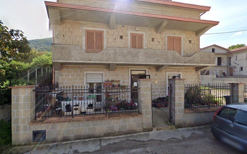 Casa in pietra a due piani con balcone e patio al piano terra, chiusa da una recinzione metallica nera. Fiori e piante in vaso decorano il patio. Un'auto grigia è parcheggiata di fronte.