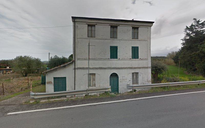 Edificio bianco a tre piani con persiane e porte verdi, adiacente a una strada rurale con guardrail metallici. Vegetazione circostante e cielo coperto visibili sullo sfondo.