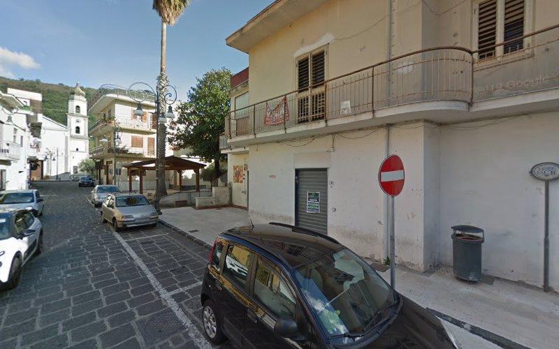 Veduta stradale di una cittadina con auto parcheggiate, un cartello di divieto di accesso, un edificio bianco con un balcone e una chiesa con due torri sullo sfondo.