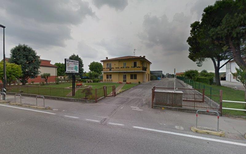 Una casa gialla a due piani con un cortile recintato è arretrata rispetto a una strada asfaltata. Il cielo è coperto di nuvole. Un cartello e degli alberi sono visibili sulla sinistra.