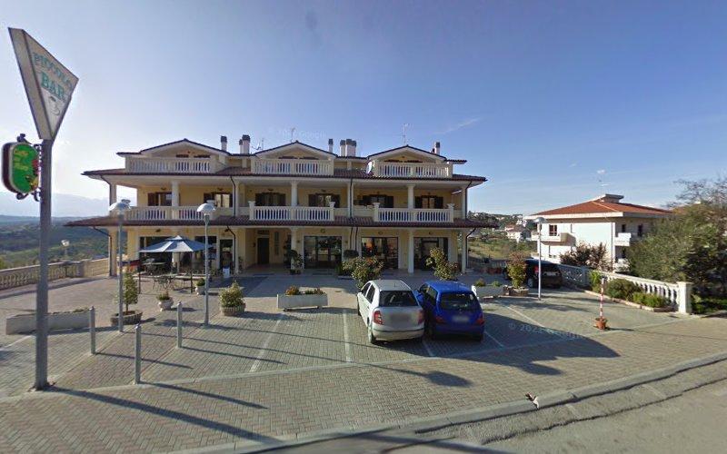 Un edificio a due piani con balconi, attività commerciali al piano terra e un parcheggio con alcune auto di fronte. L'area circostante comprende altri edifici e spazi aperti sotto un cielo limpido.
