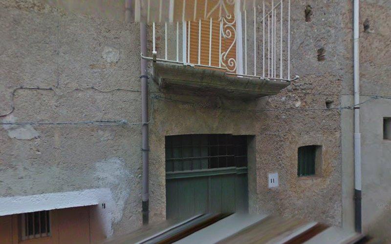 Una vecchia facciata in pietra con un piccolo balcone con ringhiere in metallo bianco sopra una grande porta verde e una piccola finestra. Le pareti sono ruvide e usurate, con fili che le attraversano.