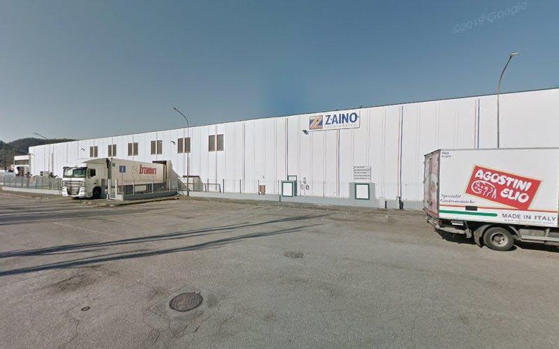 Un grande edificio industriale bianco con la scritta "Zaino". Due camion sono parcheggiati di fronte, uno con la scritta "Bram" e l'altro "Agostini Eliio - Made in Italy". La strada è vuota e pulita.
