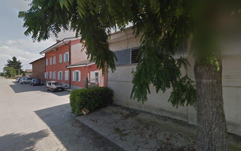 Un albero ombreggia un marciapiede accanto a un grande edificio rosso a due piani con finiture bianche e un edificio grigio. Auto parcheggiate e cespugli sono visibili sullo sfondo.