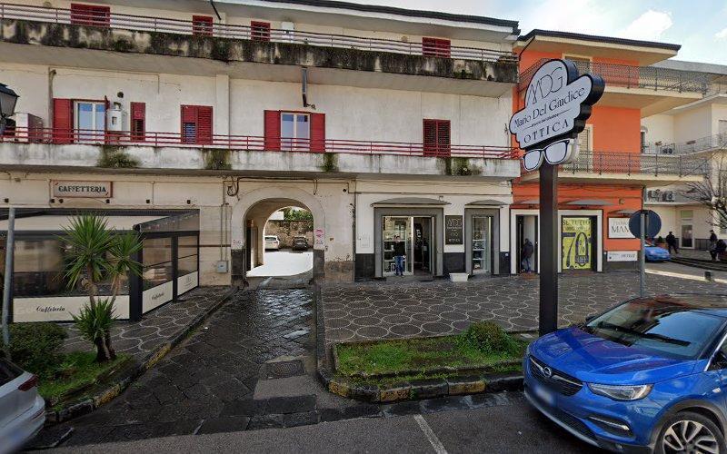 Una vista stradale mostra un edificio con persiane rosse e diversi negozi, tra cui un ottico e un bar, di fronte a un marciapiede pavimentato. Un'auto blu è parcheggiata lì vicino.