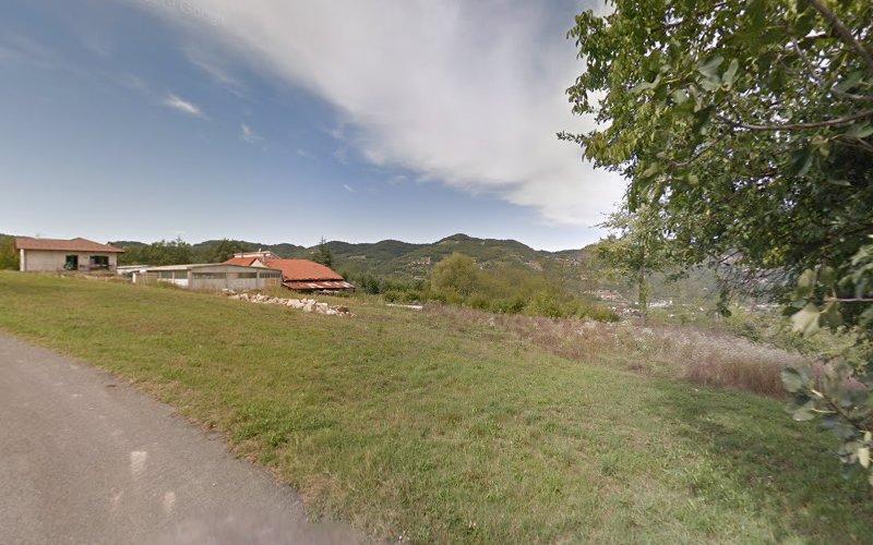 Una zona rurale con una strada stretta, campi erbosi, alcuni edifici e montagne sullo sfondo sotto un cielo parzialmente nuvoloso.