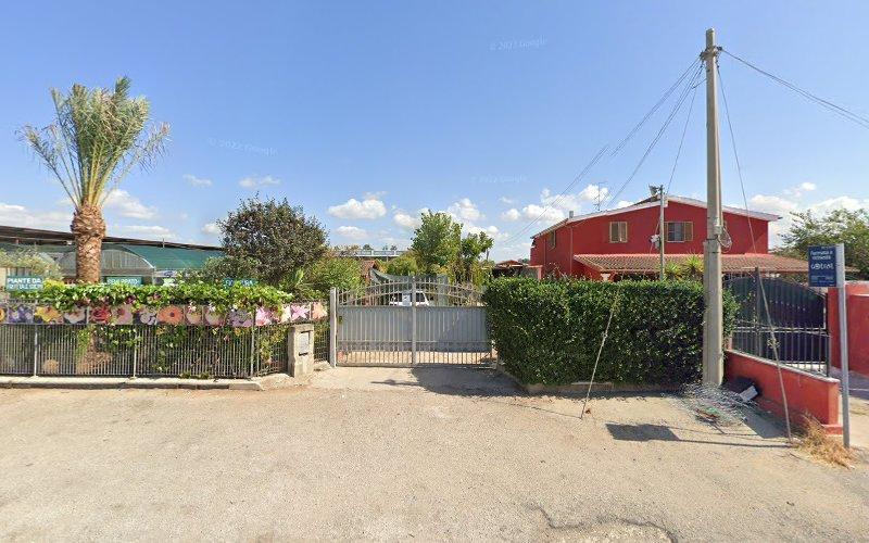 Un ingresso recintato a una proprietà con un edificio rosso e un'alta siepe. Una palma e un giardino sono visibili sulla sinistra. Un palo della luce con cavi si trova sulla destra. Cielo azzurro con nuvole sparse in alto.