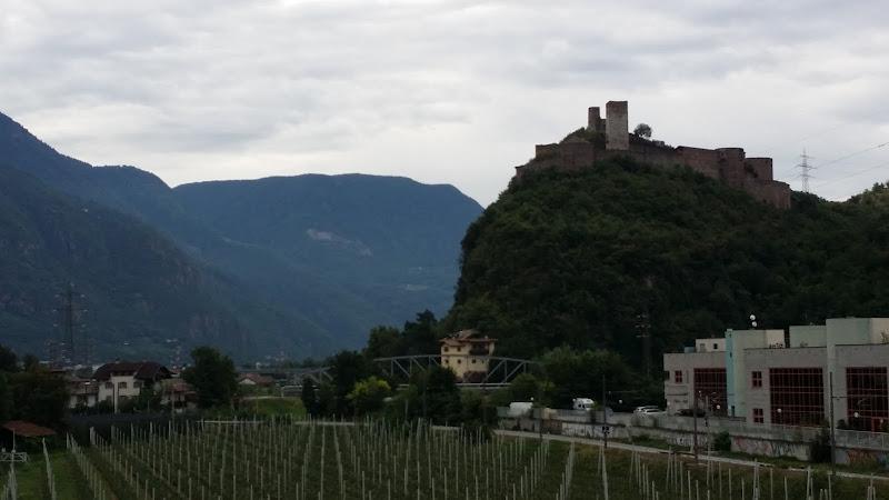 Un castello in cima a una collina domina una piccola cittadina circondata da vigneti ed edifici industriali, sullo sfondo di un terreno montuoso e di un cielo nuvoloso.