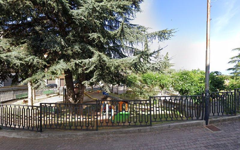 Un piccolo parco recintato con un parco giochi per bambini con scivoli e attrezzature per arrampicarsi è circondato da grandi alberi frondosi. Un sentiero pavimentato costeggia il perimetro del parco, delimitato da una recinzione nera.