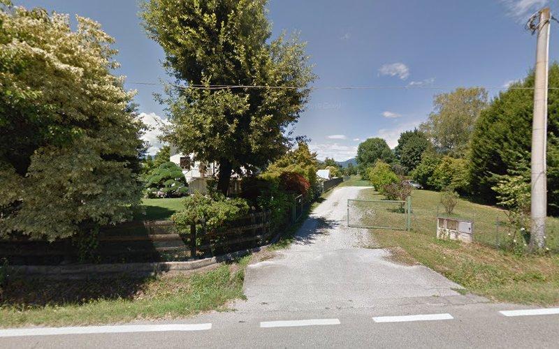 Un vialetto asfaltato conduce a una proprietà residenziale, fiancheggiata da alberi e vegetazione, con una staccionata in legno e un cancello sulla destra. Un palo della luce si trova all'ingresso. Il cielo è limpido con qualche nuvola.