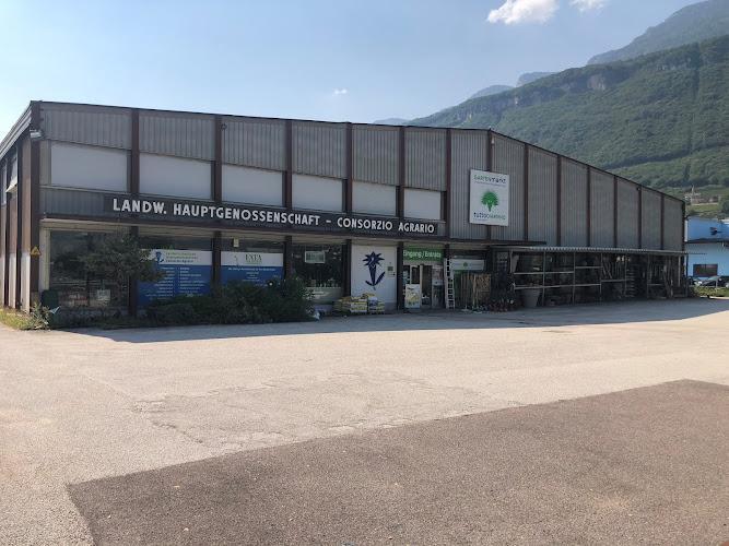 Un grande edificio rettangolare con la scritta "Landw. Hauptgenossenschaft - Consorzio Agrario" e "Agraria". L'area antistante è un ampio marciapiede vuoto con montagne visibili sullo sfondo.